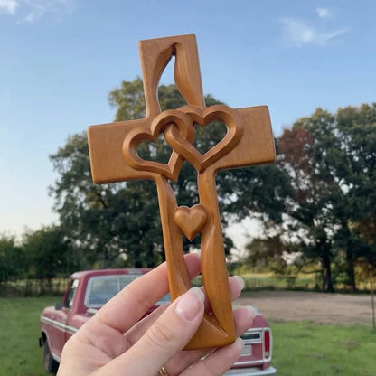Hand-carved wooden cross with two interlocking hearts, held up outdoors on a rural property